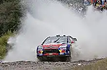 Citroën C4 WRC de Sébastien Loeb, livrée rouge et bleue, vue de face lors d'un passage de gué avec larges éclaboussures d'eau en arrière-plan.