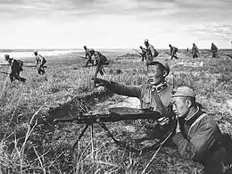 Photographie d'un soldat épaulant un fusil-mitrailleur depuis une tranchée. À l'arrière-plan des fantassins avancent prudemment dans un paysage de steppe.