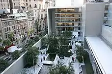 Vue de hauteur de la cour d'un bâtiment moderne, avec des arbres, des statues, des bassins (centre et droite), et de la rue adjacente (gauche).