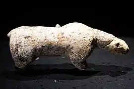 Photographie en couleurs sur fond noir et vue du dessus d'une statuette en ivoire représentant un ours des cavernes, ses pattes amputées reposant sur un socle de verre.