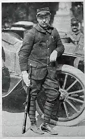 Photo d'un homme en uniforme tenant son arme et appuyé sur le passage de roue d'une voiture.