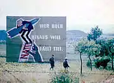 Deux soldats est-allemands photographiés à travers une clôture barbelée marchant dans un champ. Derrière eux se trouve un grand panneau de propagande représentant une caricature du chancelier est-allemand Konrad Adenauer tenant fermement un missile alors qu'il est poussé au sommet d'une échelle par un miliaire. Les barreaux de l'échelle sont formés des lettres de l'acronyme « NATO ». Le panneau porte l'inscription : « Wer hoch hinaus will, fällt tief! »