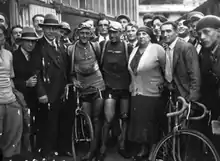 Photographie en noir et blanc montrant des cyclistes à l'arrivée d'une épreuve posant aux côtés de personnes habillées en civil.