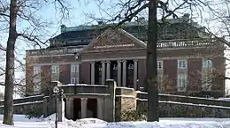 Photo du bâtiment principal de l'Académie royale des sciences de Suède, prise de face. Le bâtiment est en briques rouges et le nom de l'Académie est inscrit sur son frontispice. Deux rampes mènent à l'entrée principale, constituée de grandes portes vitrées et délimitée par des colonnes blanches.