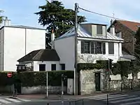 Photo en couleur d'une petite maison à un coin de rue, crépie de blanc et aux volets sombres.