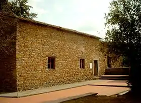 Photographie en couleurs représentant une maison en pierres ornée de deux petites fenêtres et une porte d'entrée.