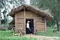 Photographie montrant un petit bâtiment en rondins de bois couvert de chaume, avec deux visiteurs à son entrée.