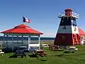 Photographie du centre d'information touristique de Grande-Anse. Le bâtiment, en forme de phare, est orné du drapeau de l'Acadie. Il y a près de celui-ci une gloriette et des tables de pique-nique peints aux couleurs du drapeau de l'Acadie. Un drapeau de l'Acadie est également monté au sommet de la gloriette.
