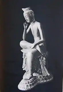 Vue de trois-quarts d'une statue assise en demi-lotus. Le pied droit repose sur la partie supérieure de la jambe gauche, le coude droit repose sur le genou droit avec la main droite près de la tête dans la pose de contemplation.