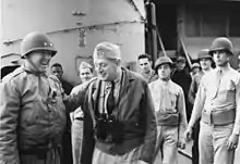 Patton portant un casque avec deux étoiles sur le devant et une veste est hilare avec un autre officier portant une veste en cuir, des jumelles et un calot. Les soldats à l'arrière-plan semblent plus circonspects.
