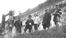 Cinq hommes à moustache – quatre militaires et un en tenue de civil (avec un document sous le bras gauche, un nœud papillon et une canne) – se tenant au-dessus d’une colline