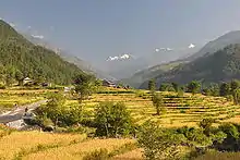 Vallée fertile avec des cultures en terrasses, quelques habitations et un passage à guet franchi par deux randonneurs, avec en arrière-plan des montagnes enneigées.