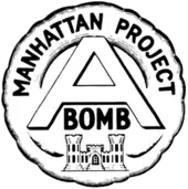 Emblème circulaire portant l'inscription « Manhattan Project » au sommet qui entoure un large « A » au centre avec le mot « bomb » écrit en dessous. L'ensemble surmonte un château qui est l'emblème du corps du génie de l'armée des États-Unis.