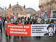 Des gens, tenant des drapeaux, des panneaux et une banderole avec notamment écrit dessus « Liberté pour Bradley Manning » en allemand, manifestent dans une rue.