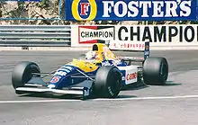 Photographie d'une Formule 1 bleue et jaune, vue de trois-quarts, sur une piste. En arrière-plan, des rails et dees panneaux publicitaires.