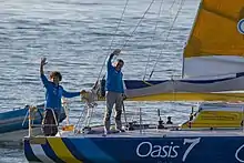 Deux hommes à l'arrière d'un voilier saluent, le bras levé.