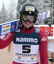 Une jeune femme en combinaison de saut à ski qui sourit avec ses skis sur l'épaule droite.