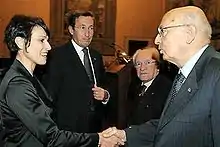 Photographie de quatre personnages en tenue de ville, une femme aux courts cheveux bruns serrant la main d'un homme dégarni portant des lunettes, deux autres hommes les regardant, en retrait.