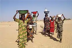 Six femmes portant des bassines d'eau portées sur leurs têtes, dans le désert