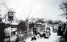 Photo noir et blanc de quelques femmes et enfants sur un chemin entre des palissades, des cabanes et un moulin derrière