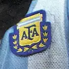 Photographie en couleurs. Gros plan sur l'écusson officiel de la fédération argentine de football cousu sur un maillot bleu ciel et blanc.