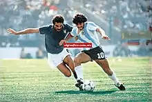 Photographie en couleurs. Un joueur italien, Gentile, à gauche avec son maillot bleu foncé, est aux prises avec un joueur argentin, Maradona, à droite avec son maillot rayé bleu ciel et blanc.