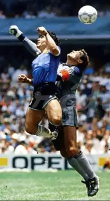 Photographie en couleurs. Deux joueurs sont en suspension pour atteindre un ballon au-dessus d'eux. Tous les deux ont un bras en l'air, tandis que Maradona a sauté plus haut que Shilton.