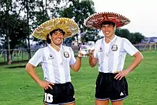 Photographie en couleurs. Diego Maradona et Daniel Passarella posent en tenue de football argentine et avec un chapeau mexicain, un verre à la main, tout sourire.