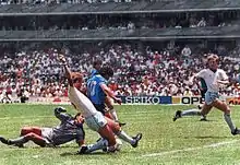 Photographie en couleurs. L'Argentin tire tandis que le gardien anglais est au sol derrière lui et suit le ballon du regard et qu'un défenseur anglais colle Maradona et amorce un tacle en retard.
