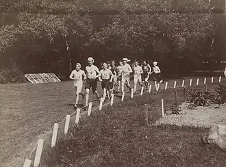 Photo en noir et blanc d'une dizaine d'hommes portants des couvre-chefs courant sur de l'herbe.