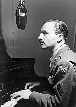 Photographie en noir et blanc d'un homme habillé en militaire et assis devant un piano et un microphone de la BBC.