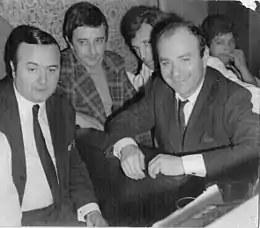 La phot noir et blanc représente des personnes assises autour d'une table de café dont Marcel Rossi et Fernand Raynaud