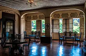 Salon en bois, avoir trois tables en bois et des chaises. Des panneaux coulissant en bois persiennes sont présents sur toute la longueur du mur extérieur.