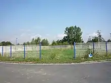 Photographie montrant un rond-point partiellement entouré de grilles blanches et envahi de mauvaises herbes, au centre duquel se trouve la tête de puits matérialisée de la seconde fosse de Marchiennes, ouverte par la Compagnie des Canonniers de Lille.