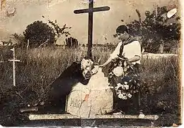 Photo en noir et blanc montrant un homme agenouillé sur une tombe ornée d'une croix, avec un autre homme debout à côté, tenant une couronne de fleurs
