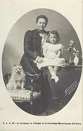  la comtesse de Flandre, portant une robe sobre et foncée, ainsi qu'un long sautoir sourit en tenant sa petite-fille en tenue claire, tandis qu'à droite est assis un loulou de Poméranie