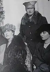 Photo noir et blanc de deux femmes assises, une femme debout en arrière-plan. Toutes trois portent un chapeau.