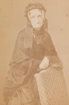 Portrait photographique d'une femme se tenant debout, appuyée sur le dossier d'une chaise, portant une coiffe et une robe noire.