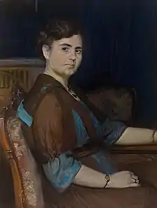 Halbfigürliches Portrait einer sitzenden Dame, den Kopf zum Betrachter gewandt, vor blauem Vorhanghintergrund. Louise Catherine Breslau.1912.