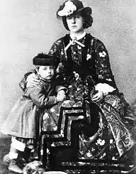Léopold âgé de cinq ans, vêtu de pantalons courts, d'une longue veste et coiffé d'une toque foncée se tient debout près de sa mère assise et vêtue d'une robe à crinoline sombre aux motifs chamarrés et coiffée d'un chapeau sombre orné d'une plume claire