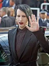 Homme de style gothique habillé d'un costume noir, au visage maquillé de blanc avec un rouge à lèvres, saluant de la main.