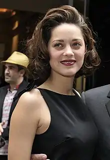  Photographie en couleurs d'une femme brune aux traits fins, vêtue d'une élégante robe noire, sur un tapis rouge.