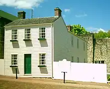 Façade coté rue de maison traditionnelle à un étage, peinte en blanc, avec haute palissade à droite.