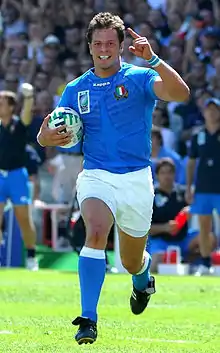Photographie d'un joueur de rugby vêtu d'un maillot bleu, d'un short blanc et portant un ballon dans la main droite
