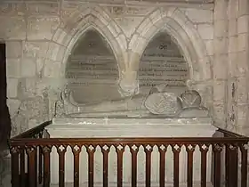 Photo d'un gisant en pierre, les mains jointes, précédé au premier plan d'une rambarde en bois. Au troisième plan un texte peint sur le mur est consacré à Enguerrand de Bournonville.