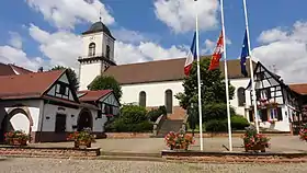 Photographie couleur d'une église surplombant une place fleuries et des maisons à colombage