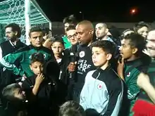 Un joueur de football parle avec un micro, à côté de dizaines de supporters, près d'un but.