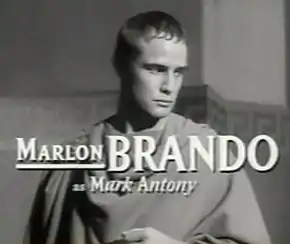 Photographie en noir et blanc d'un homme portant la toge, le regard pensif vers la bas de l'image. Des caractères de grande taille, en surimpression, indiquent que Marlon Brando joue le rôle de Marc Antoine.