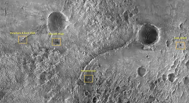 Sur cette photo, également prise par le satellite MRO, on peut distinguer les points de chute des différents composants de la sonde spatiale sur le sol martien : de gauche à droite le parachute et le bouclier thermique arrière, le point d'impact du skycrane, le rover Perseverance et le point d'impact du bouclier thermique avant.