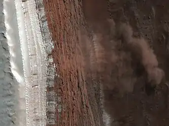 Glissement de terrain observé par l'instrument HiRISE de MRO le 19 février 2008. La glace d'eau est bien visible au sommet de la falaise sous forme d'une couche blanche. C'est une partie de ce matériau qui a glissé en contrebas, formant une « avalanche » à l'origine de volutes de poussières et de cristaux de glace mêlés suffisamment épaisses pour projeter leur ombre en dessous d'elles. Le soleil éclaire la scène depuis l'angle supérieur droit.